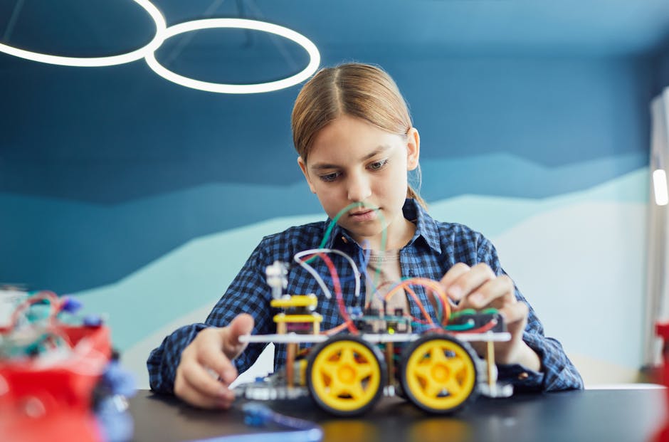 Teenage girl working on a robotic car project with wires and wheels inside a classroom.
