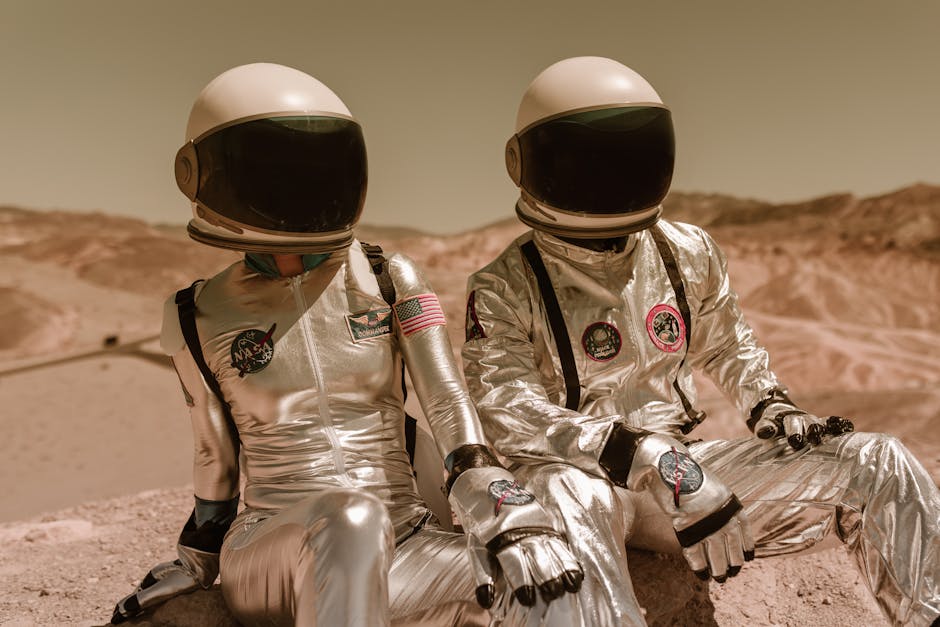 Two astronauts in silver suits and helmets sitting on a Mars-like desert landscape.